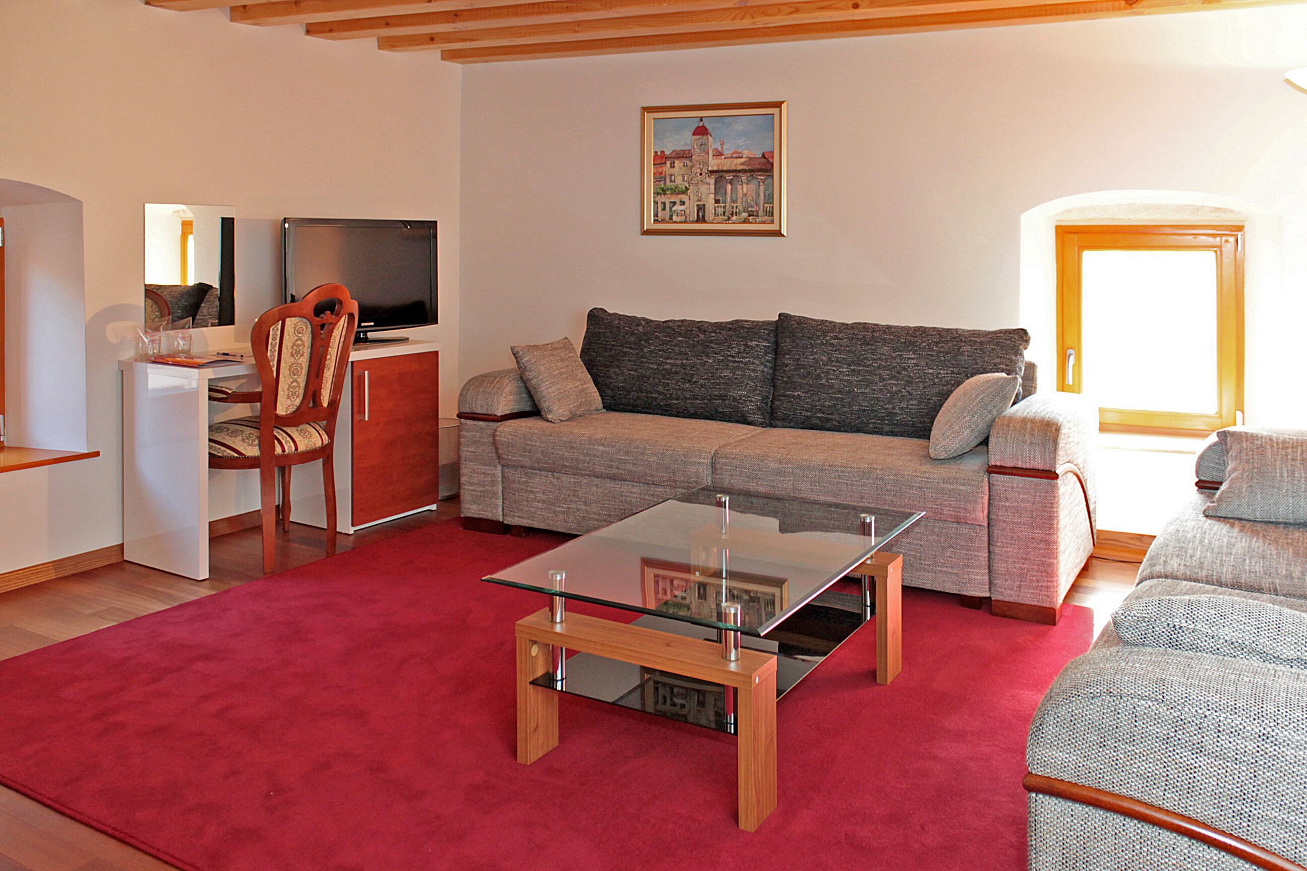 A living room with a sofa, a glass coffee table, a red carpet, a wooden desk, and a television.