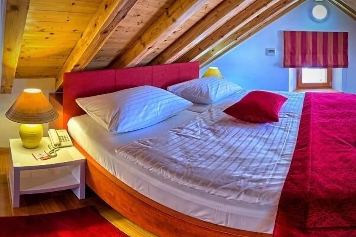 A cozy attic bedroom with a curved bed, a bedside lamp, and a window with a red valance.