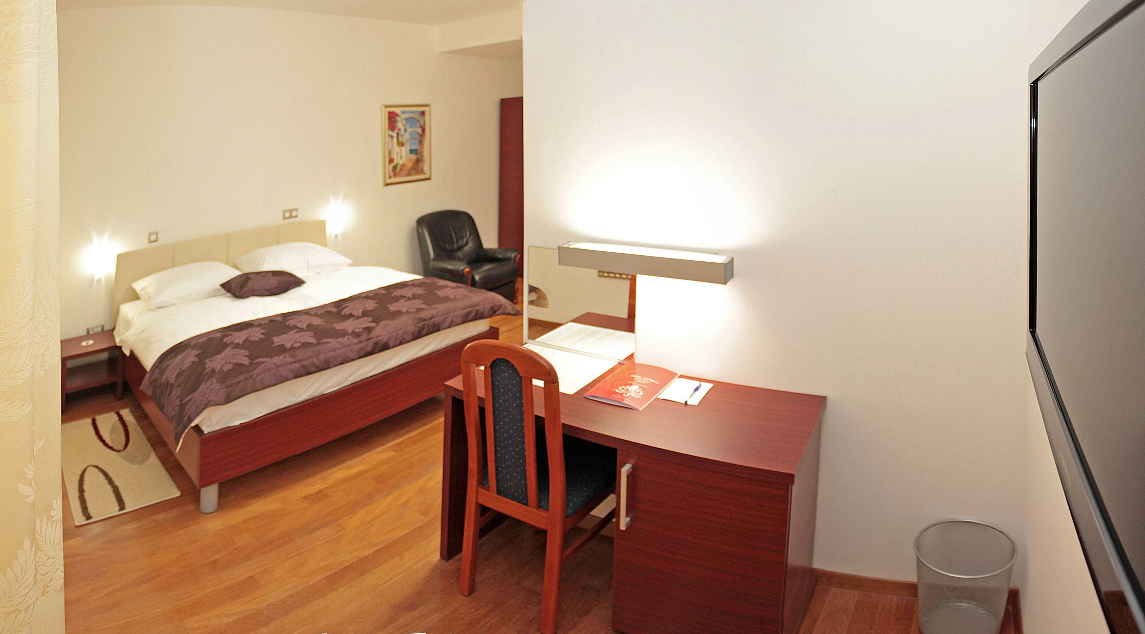 A hotel room with a bed, a desk with a chair, a lamp, and a trash bin.