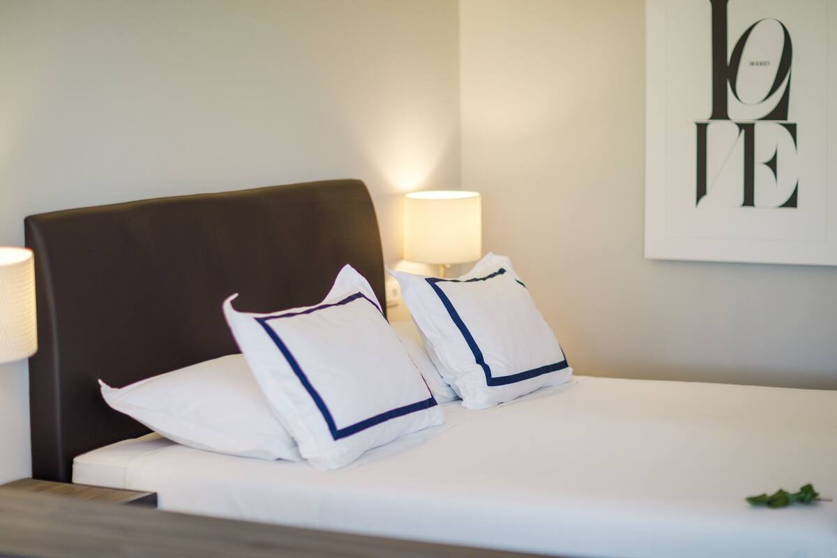 Een opgemaakt bed met witte lakens en marineblauwe kussens, een donker hoofdbord en een muur met een 'LOVE'-bord.