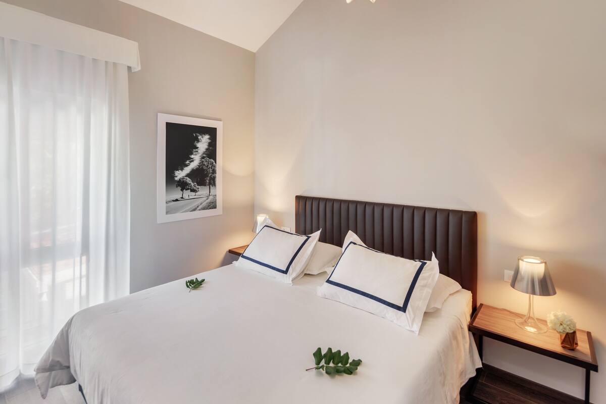 Een netjes opgemaakt bed met witte lakens en blauwe accenten, een nachtkastje met een lamp, en een ingelijst schilderij aan de muur.