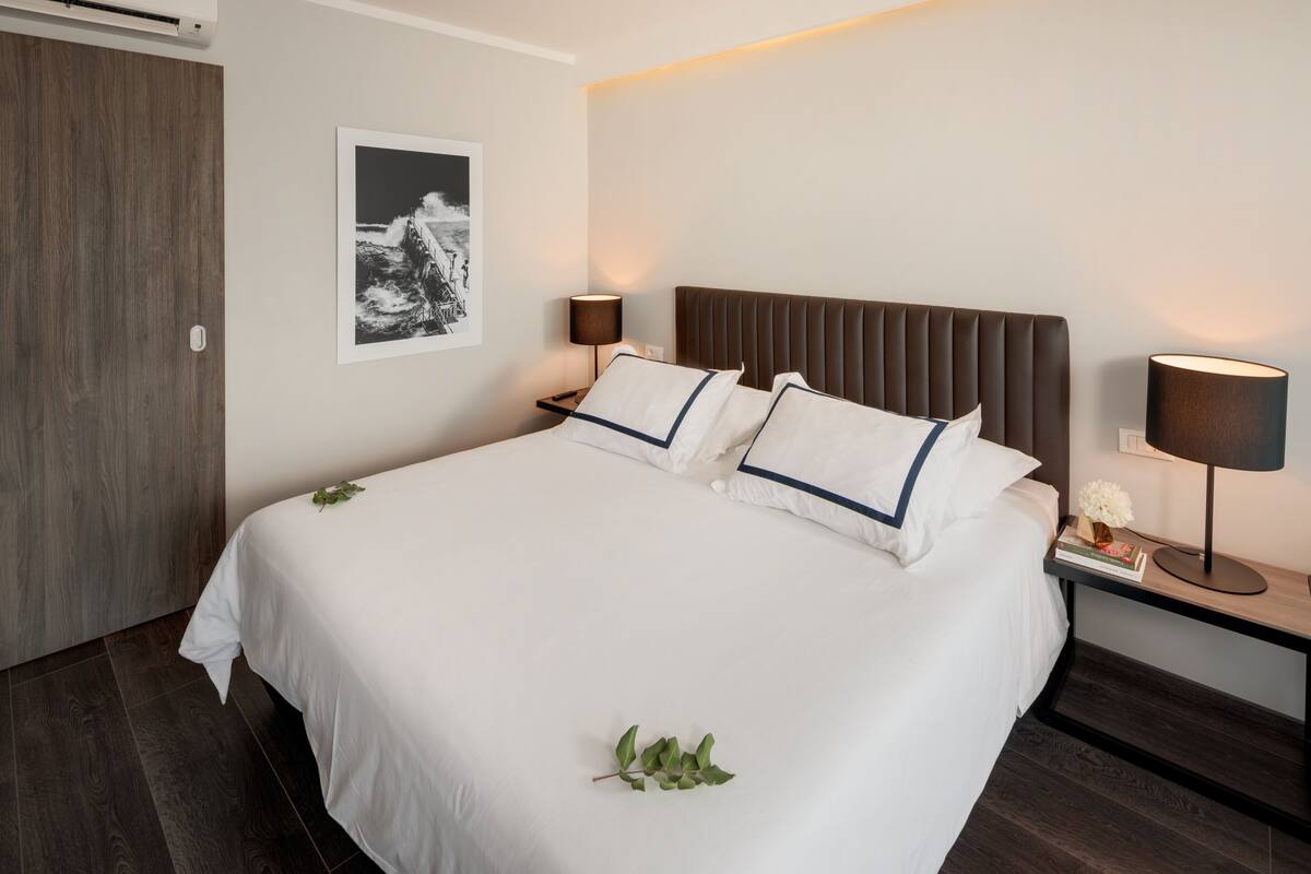Een netjes opgemaakt bed met witte lakens en blauwe accenten, een nachtkastje met een lamp, een ingelijst schilderij aan de muur en een houten deur.