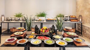 Een buffetopstelling met diverse gerechten en dranken, waaronder fruit, salades en desserts, gepresenteerd op tafels met potplanten en kaarsen.