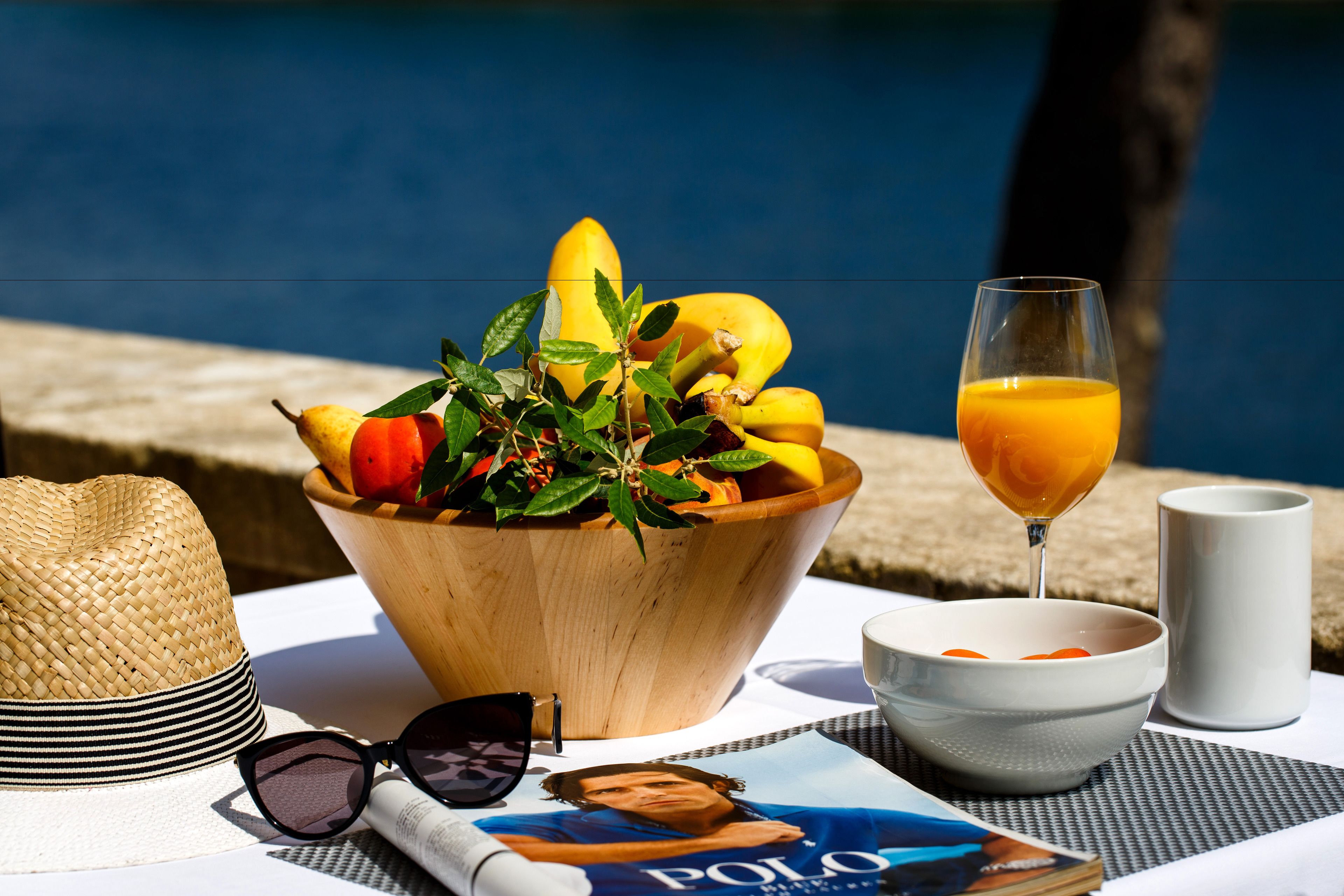 Een houten schaal met fruit, een glas sinaasappelsap, een strohoed, zonnebril en een tijdschrift op een tafel bij een zwembad.