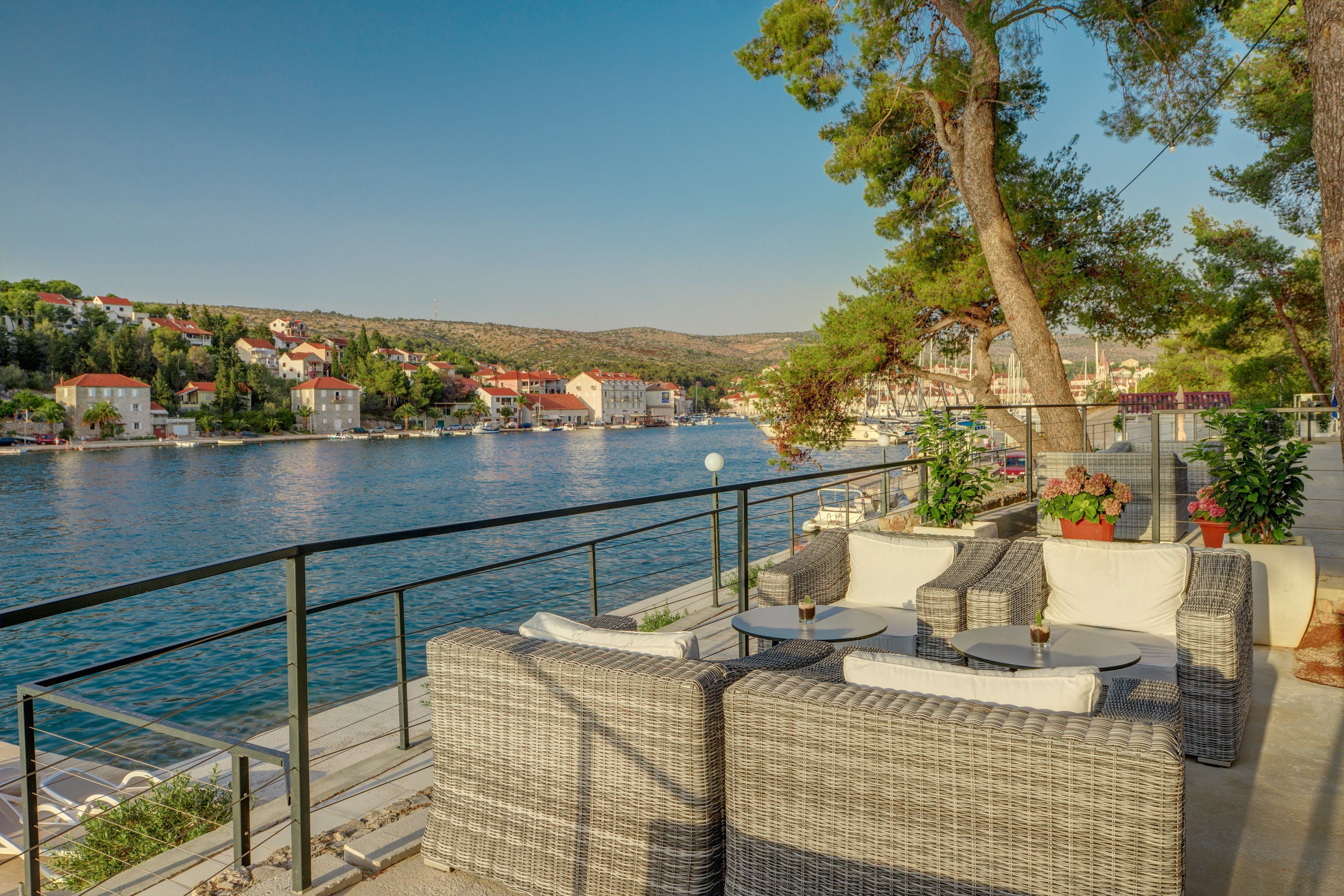 Une terrasse au bord de l’eau, meublée de mobilier en osier, offrant une vue sur la mer et les bâtiments.