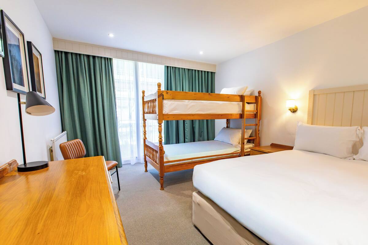 Quarto de hotel com beliche, escrivaninha, cadeira, abajur e janela com cortinas.