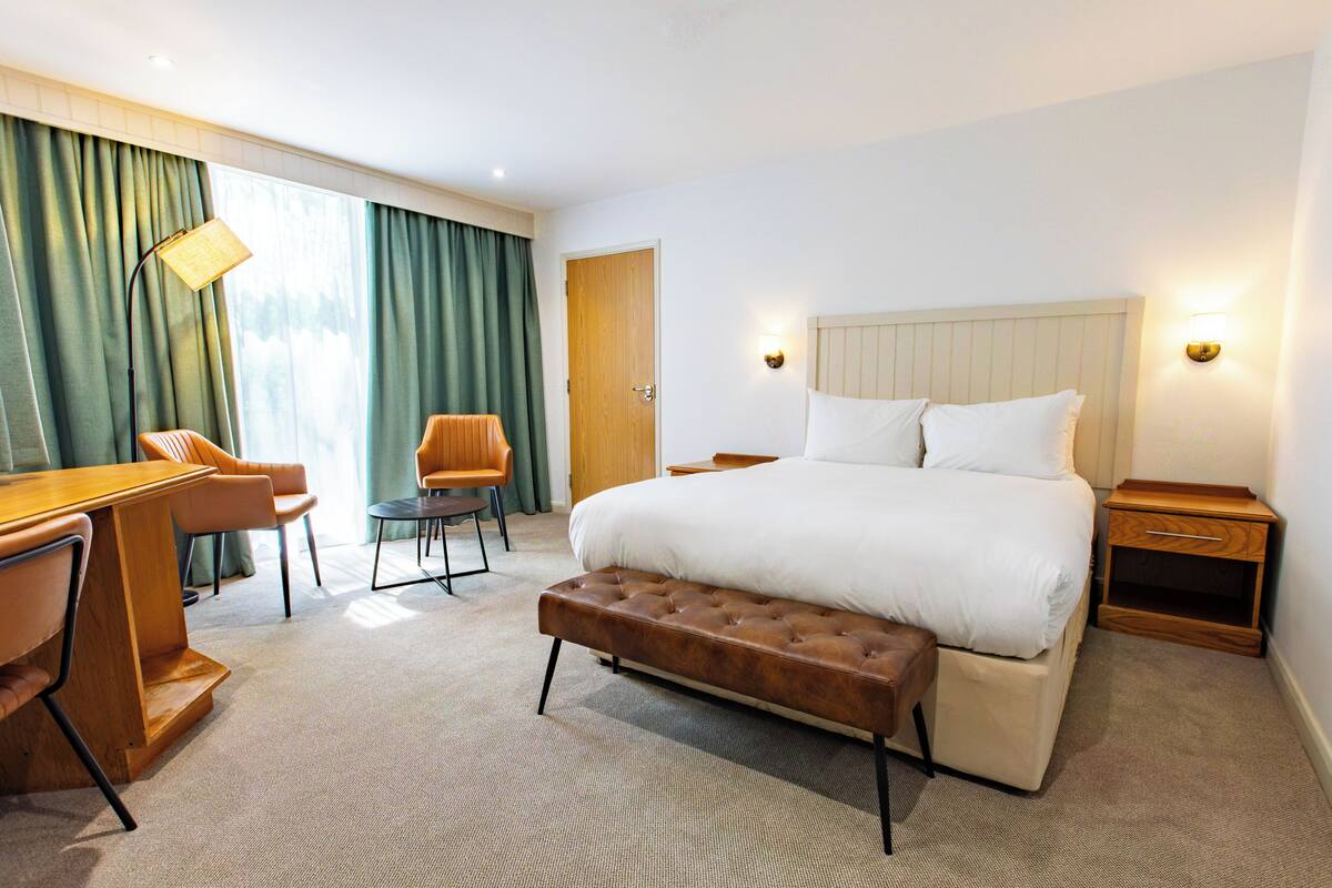 Quarto de hotel com uma cama grande, uma escrivaninha com cadeira, uma mesinha, um banco, uma porta e uma janela com cortinas.