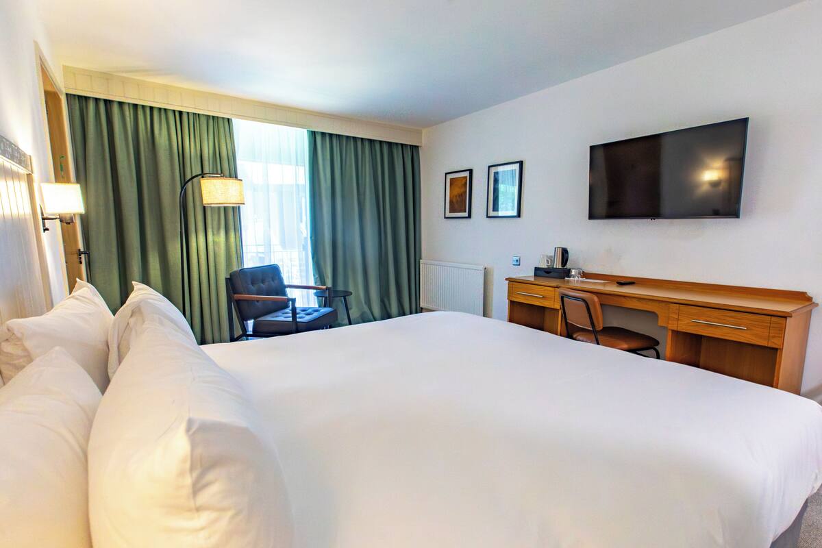 Quarto de hotel com cama grande, escrivaninha, cadeira, televisão e janela com cortinas.