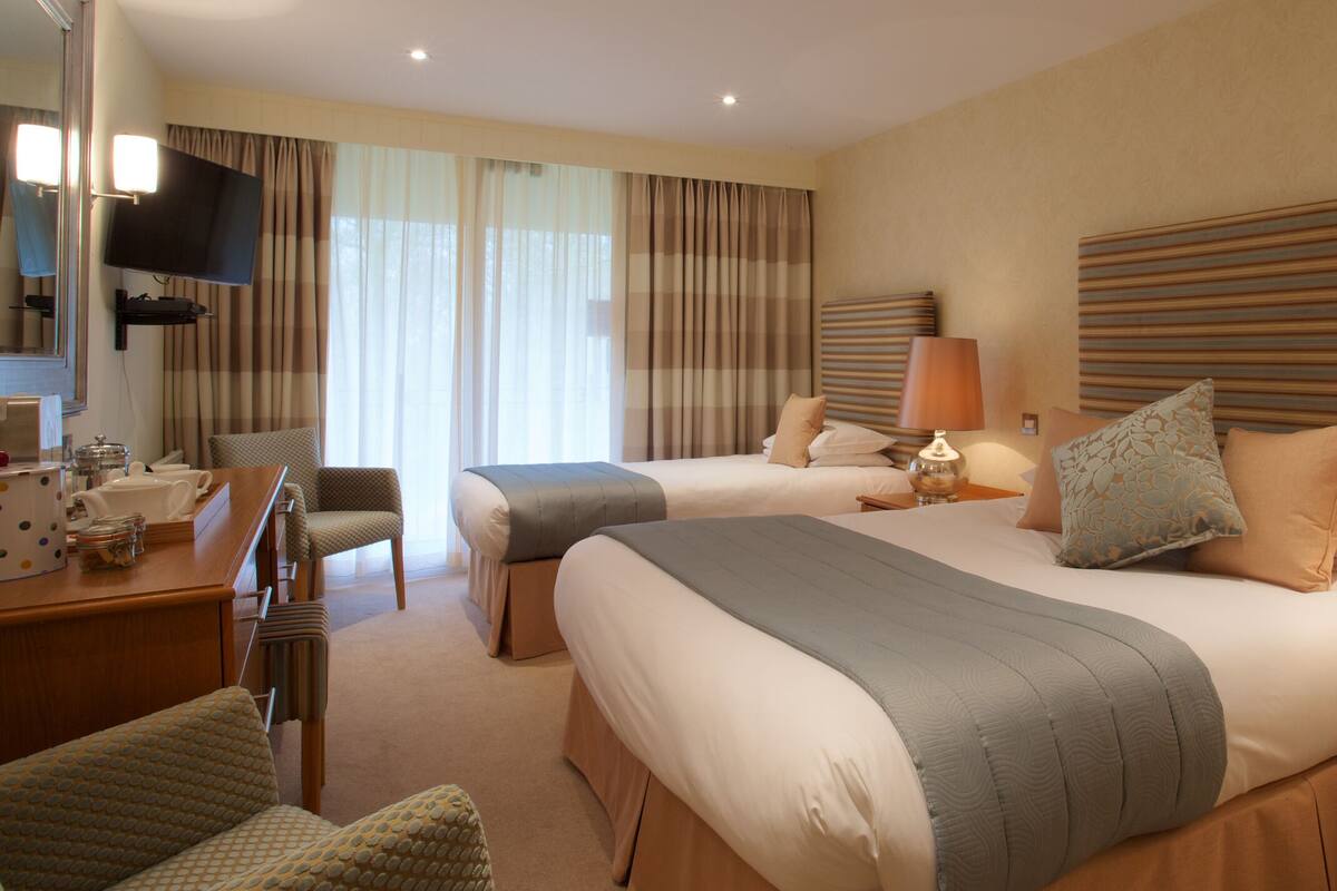 Quarto de hotel com duas camas, televisão, escrivaninha com cadeira e uma janela grande com cortinas.