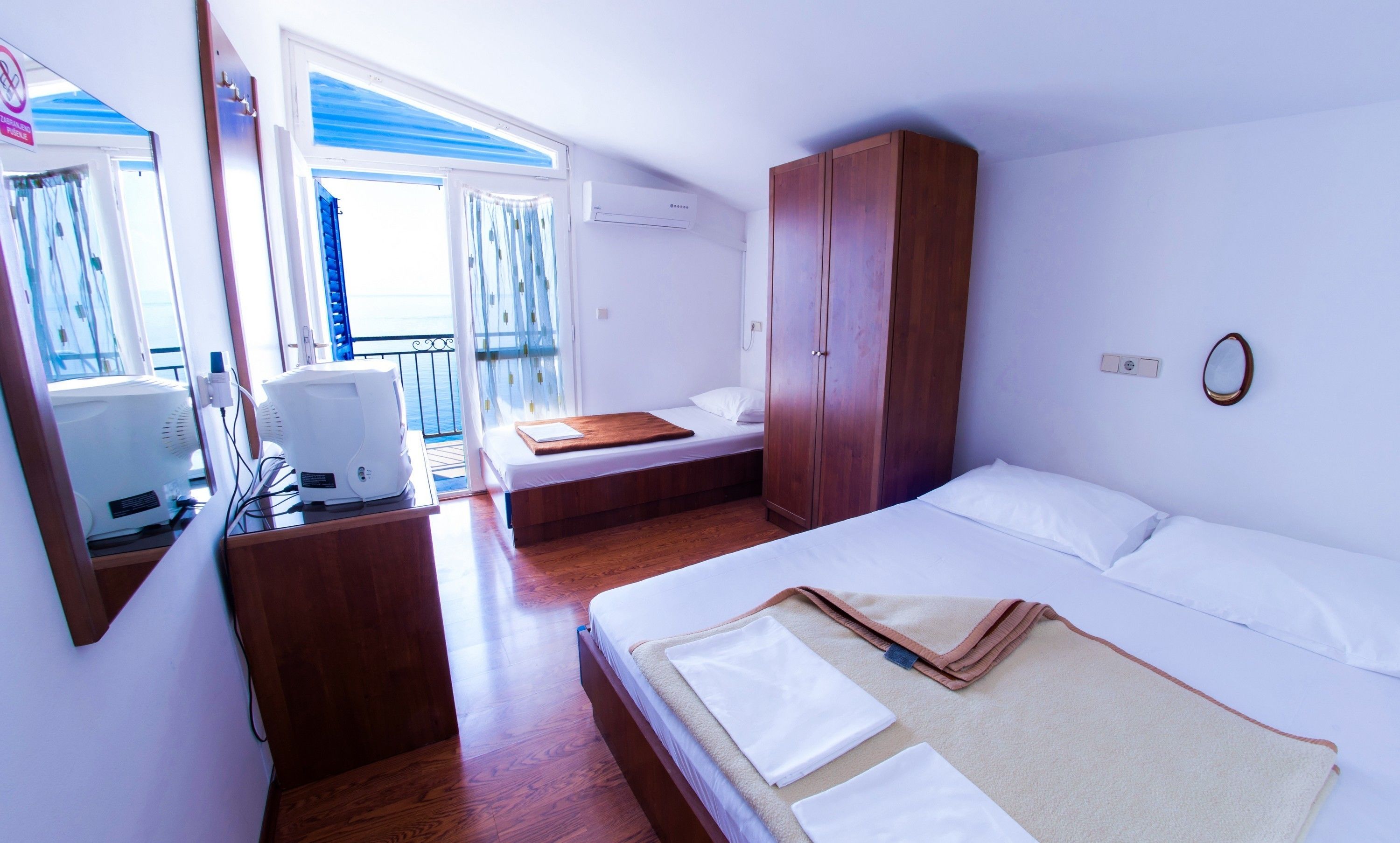 A hotel room with a bed, a desk with a computer, a wardrobe, and a view of the sea through a balcony.