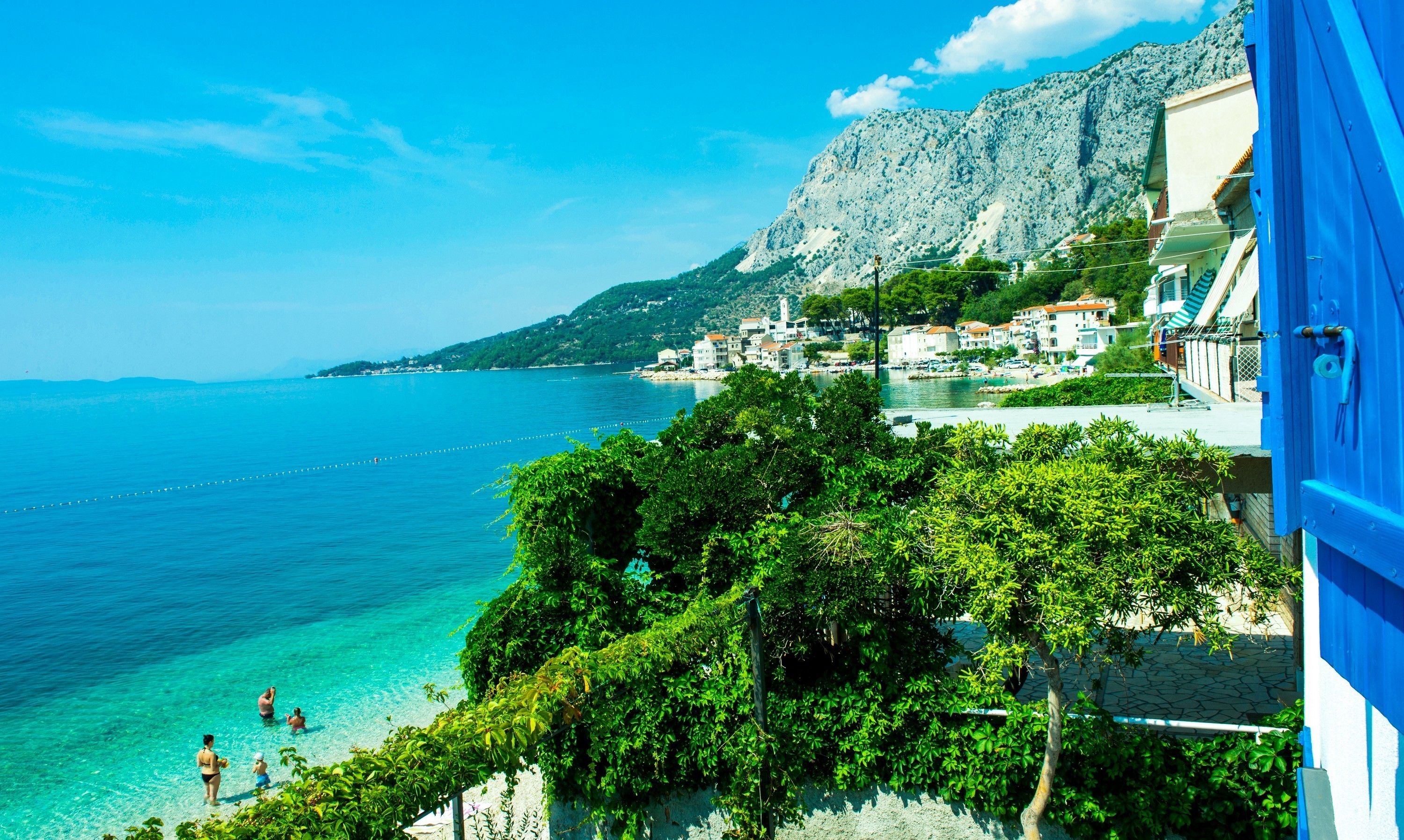 A coastal town with buildings along the sea, a prominent mountain in the background, and clear blue waters.