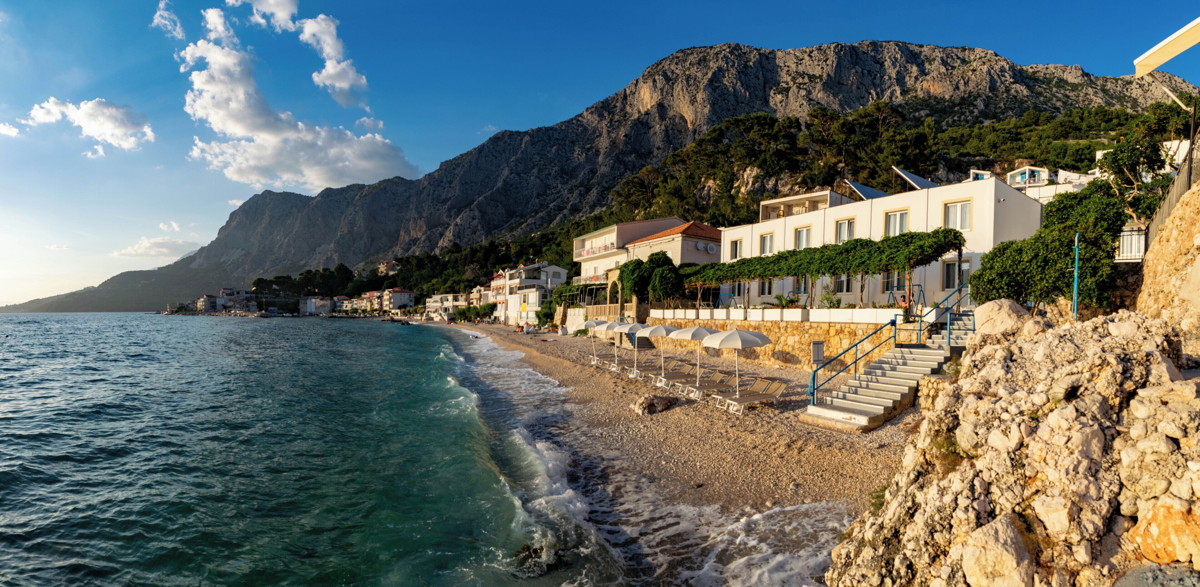A coastal hotel with a rocky beach, a mountain backdrop, and a clear sky.