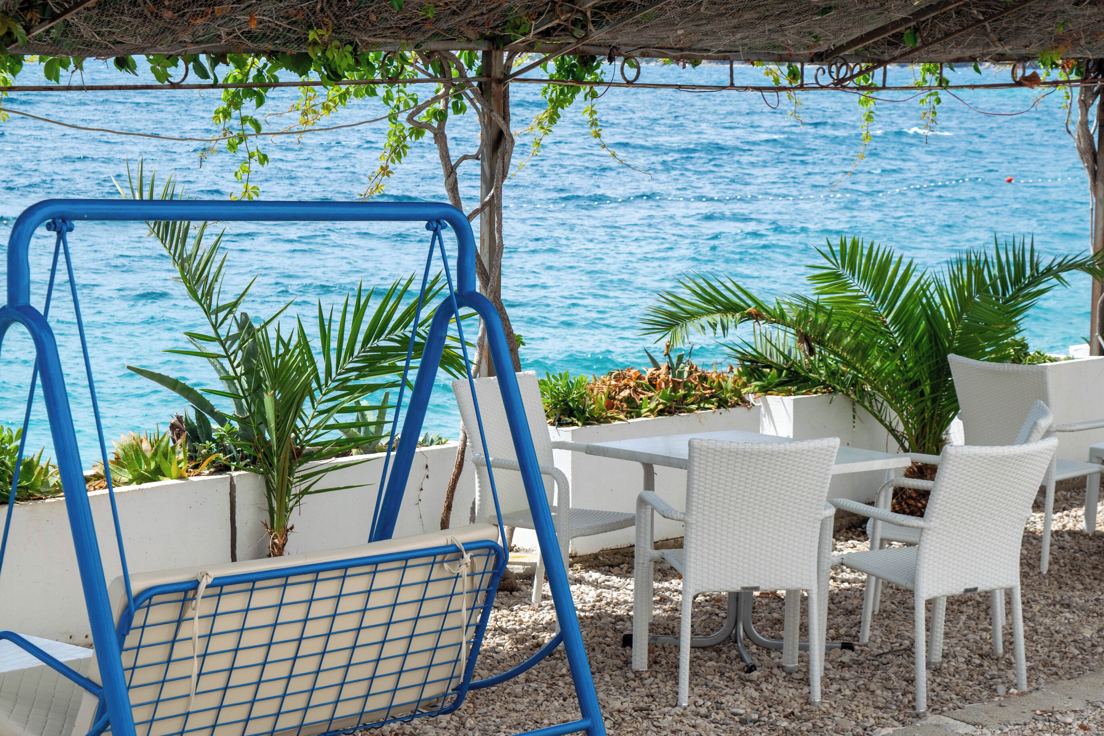 A beachside seating area with a swing chair, white wicker chairs, and a table, overlooking the sea.