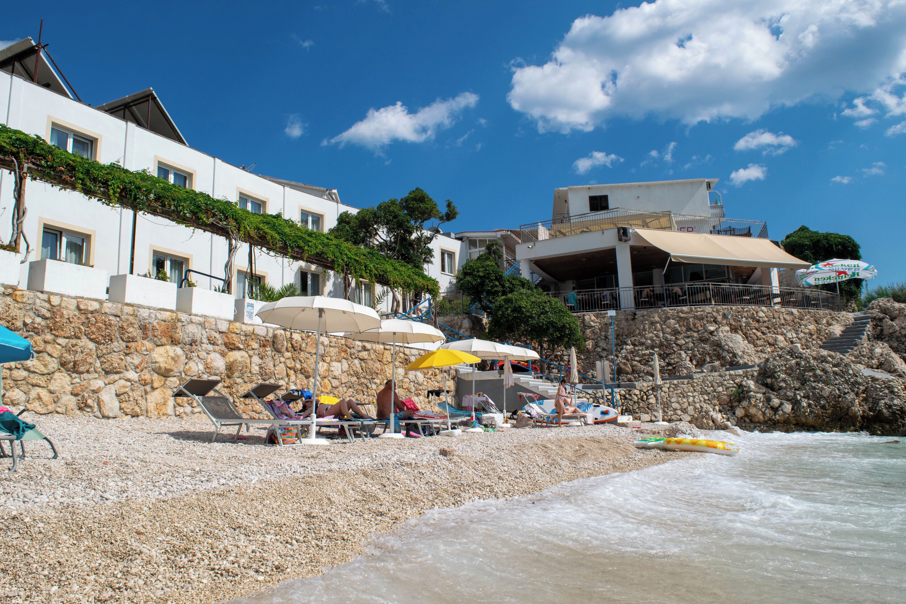 A beach with sun loungers and umbrellas, a stone wall, and a building with a balcony overlooking the sea.