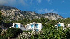A coastal building with a white exterior and blue shutters, surrounded by lush greenery and mountains in the background. - Beach Hotel Plaza (Podgora)