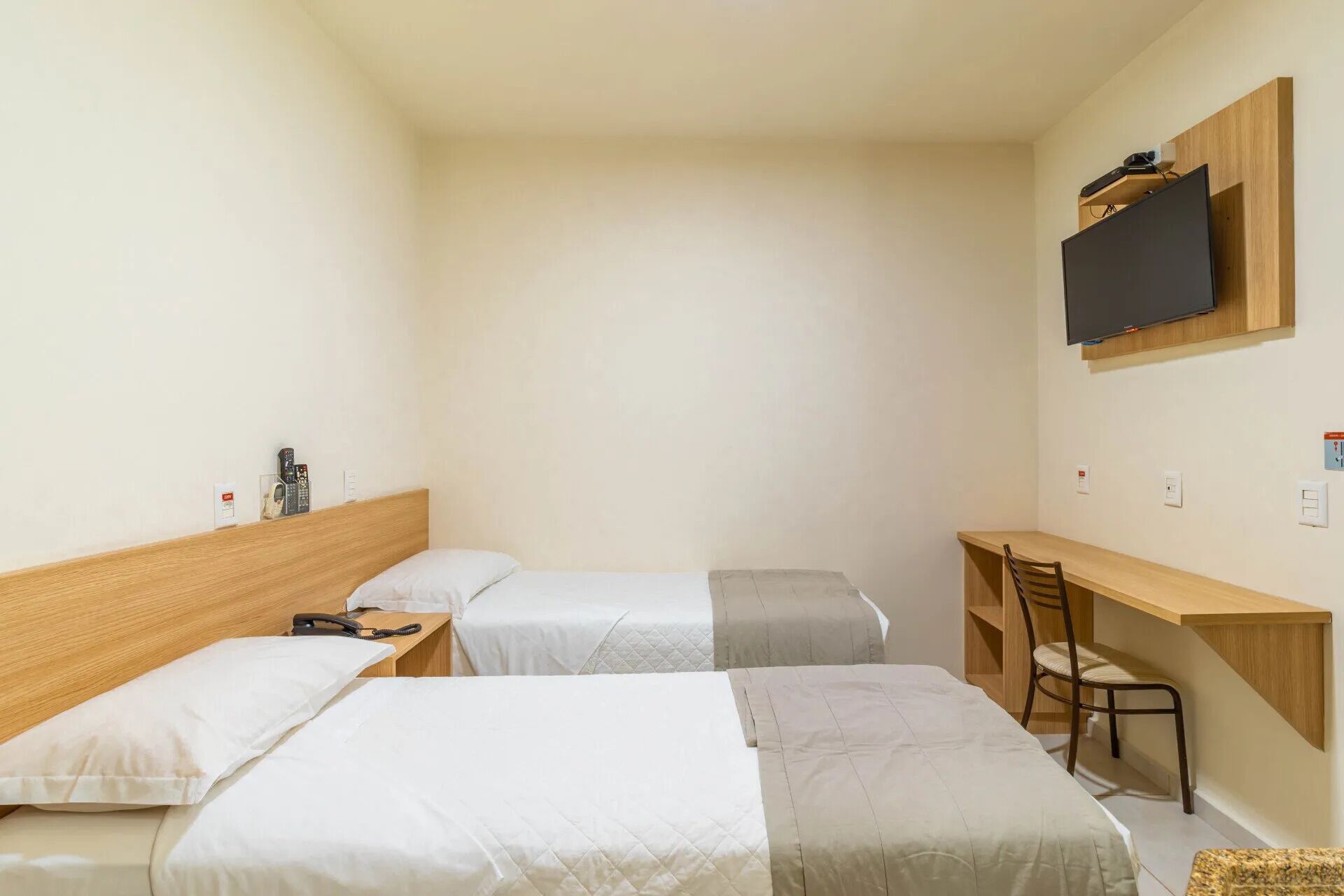 Une petite pièce bien éclairée, équipée de deux lits simples, d’un bureau, d’une chaise et d’un téléviseur fixé au mur.