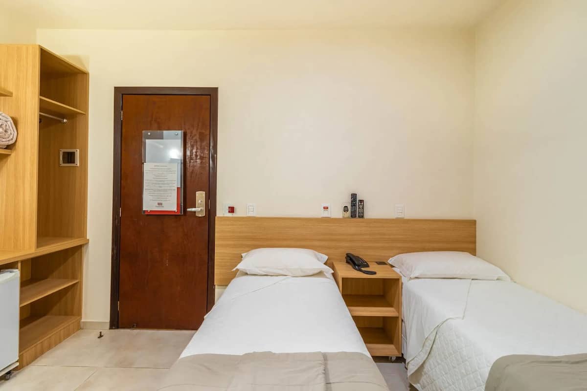 A hotel room with two single beds, a wooden wardrobe, a desk with a telephone, and a small refrigerator.