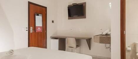 A hotel room with a bed, a desk, a chair, a television mounted on the wall, and a bathroom visible through an open door.