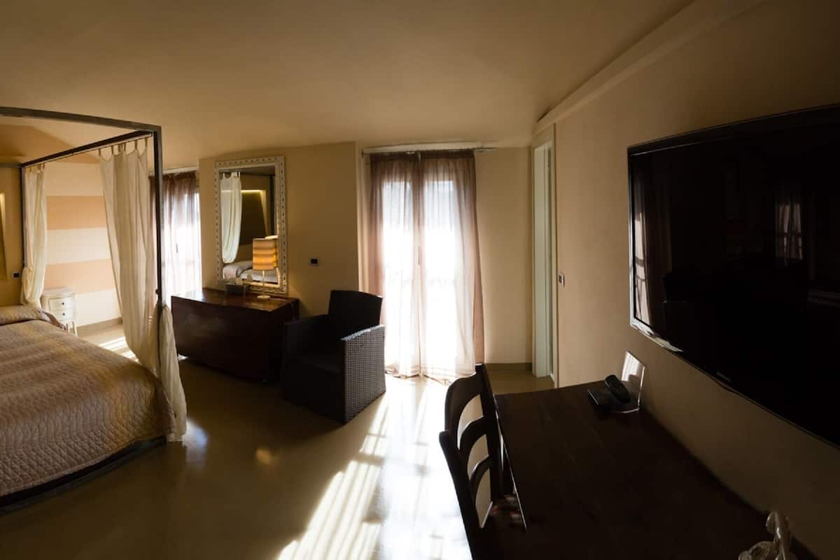 A hotel room with a four-poster bed, a sitting area with a sofa and a table, a large flat-screen TV, a bathroom with a bathtub, and a large mirror.