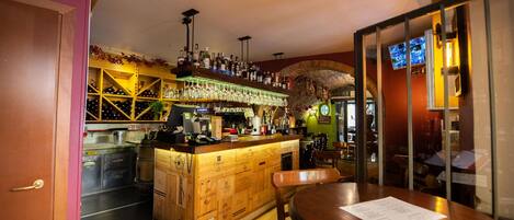A bar area with wooden bar counter, shelves with bottles, and a seating area with a table and chairs.