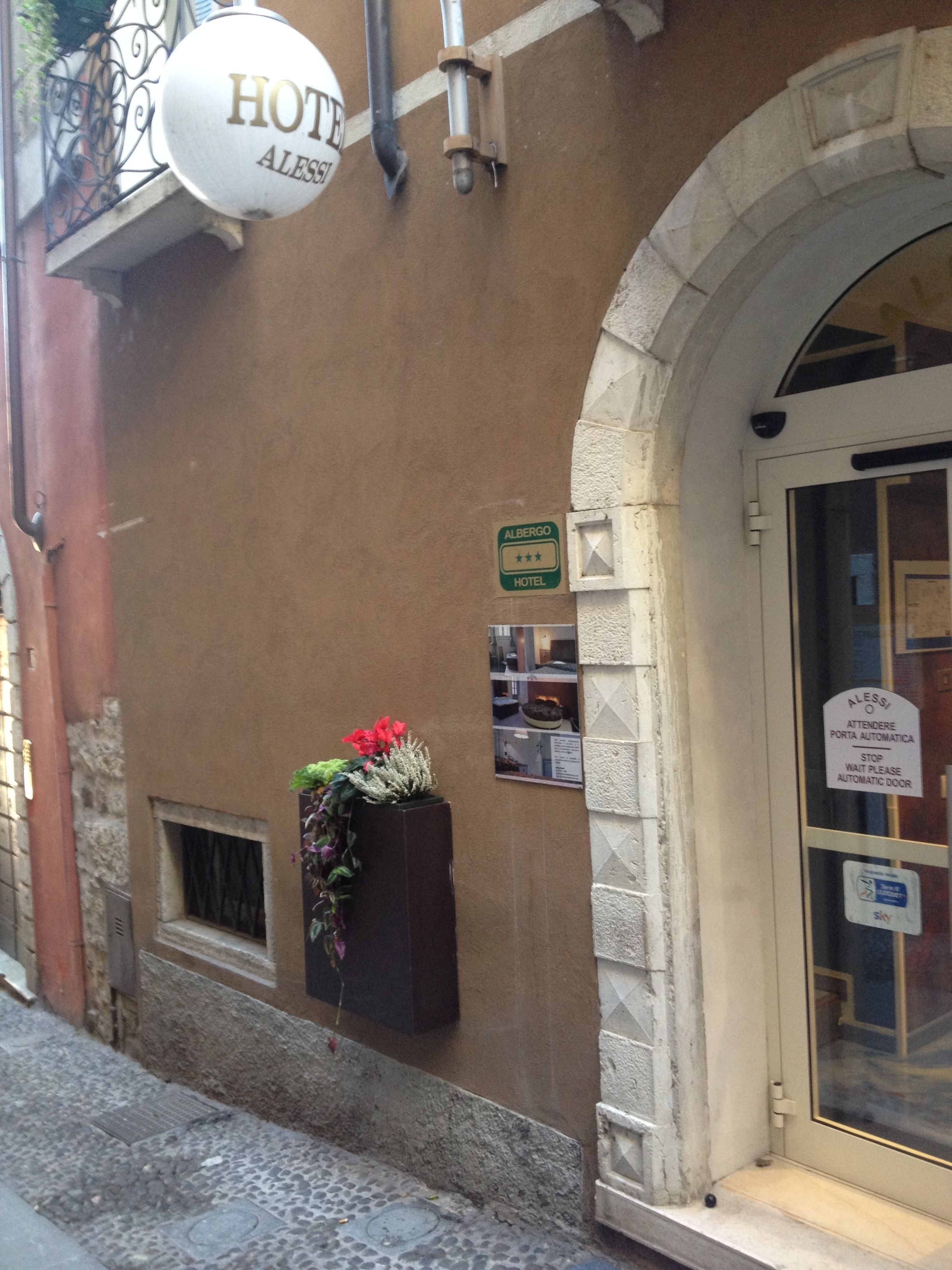 L'ingresso di un hotel con una segnaletica che indica "HOTEL ALESSI", una fioriera e un'insegna per porta automatica.