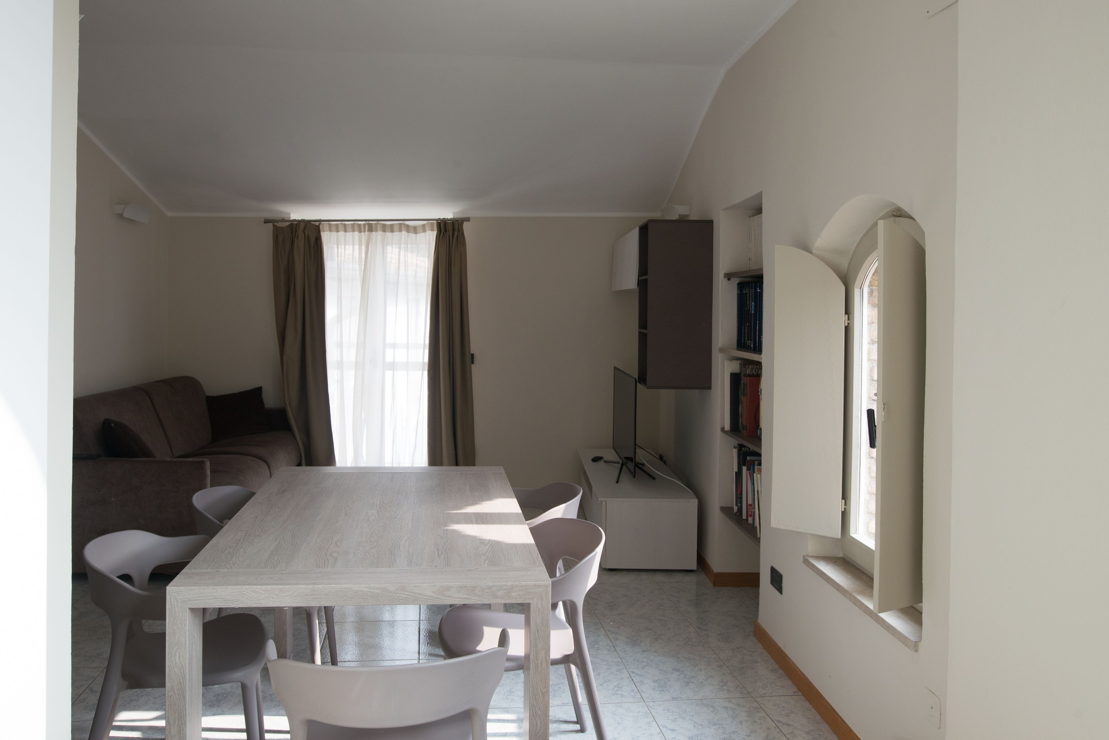 A modern living room with a dining table, white chairs, a sofa, and a bookshelf.
