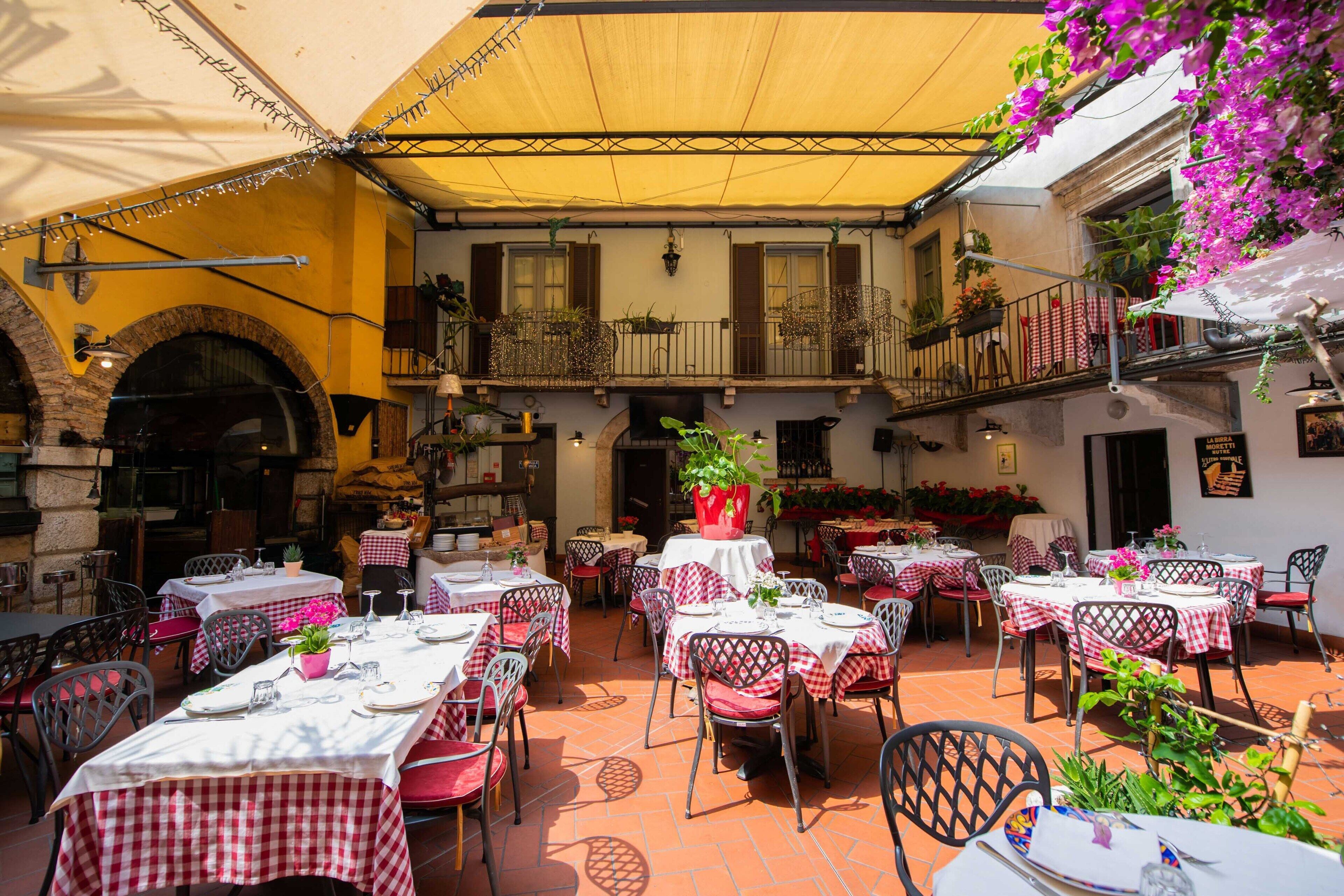 An outdoor dining area with tables set for two, covered by a yellow canopy, and surrounded by potted plants and flowers.