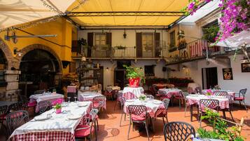 An outdoor dining area with tables set for two, covered by a yellow canopy, and surrounded by potted plants and flowers.