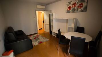 En kompakt opholdsstue med en sofa, et spisebord med stole, et lille køkken og en dør, der fører til et andet rum.