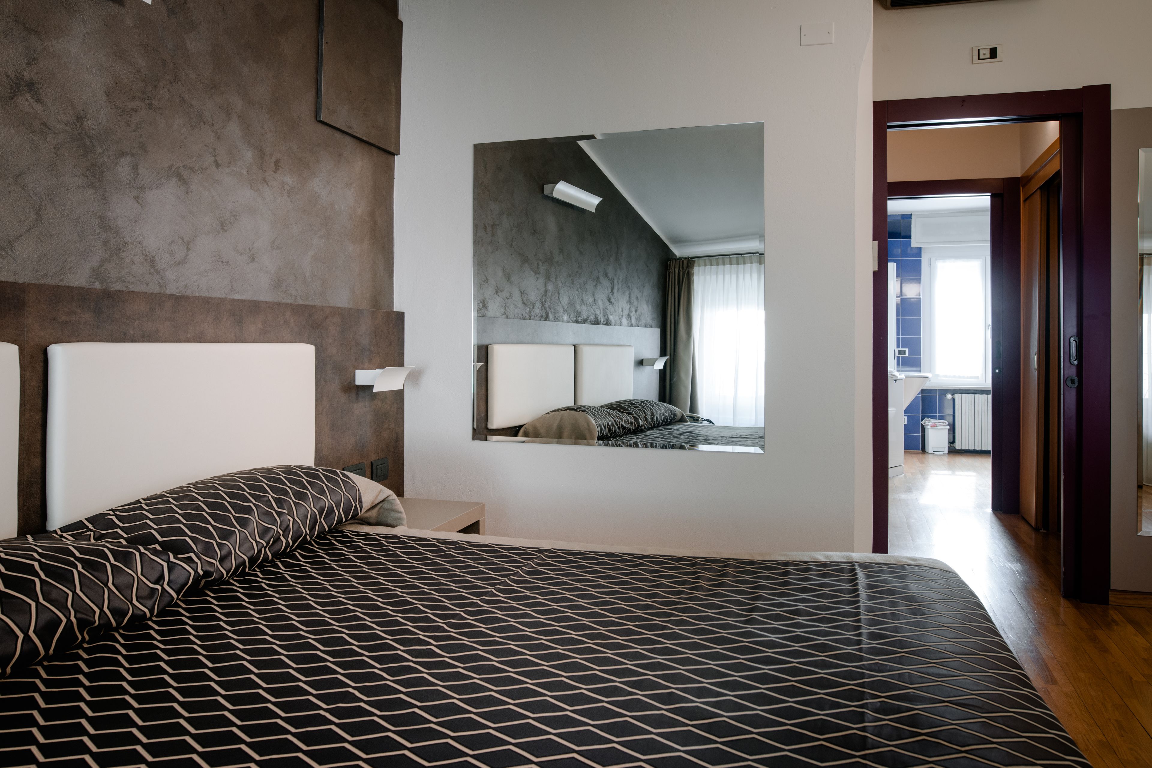 Una habitación de hotel moderna con una cama grande, cabecero integrado, mesita de noche pequeña y un espejo amplio.