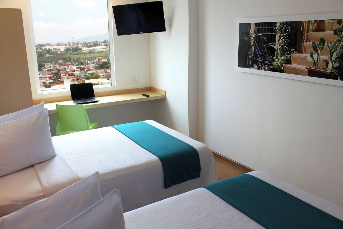 Habitación de hotel con dos camas, un escritorio con una computadora portátil, un televisor montado en la pared y una ventana con vista a la ciudad.