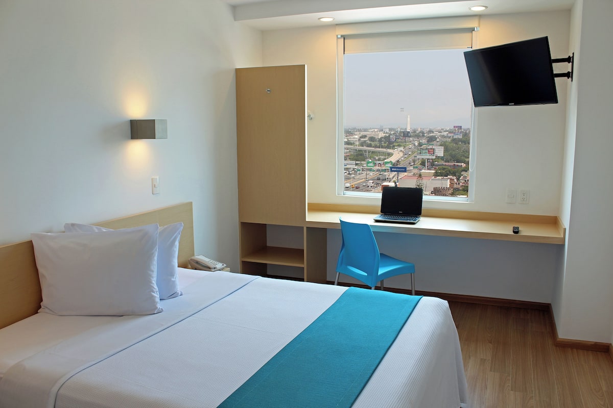 Una habitación de hotel con una cama, un escritorio con una computadora portátil, una silla, un televisor montado en la pared y una ventana con vista a la ciudad.
