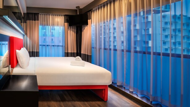 Una camera d'albergo moderna con un letto grande, una televisione a schermo piatto e vista sulla città attraverso tende trasparenti.