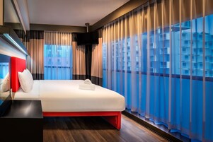 Una habitación de hotel moderna con una cama grande, un televisor de pantalla plana y vistas a la ciudad a través de cortinas translúcidas.