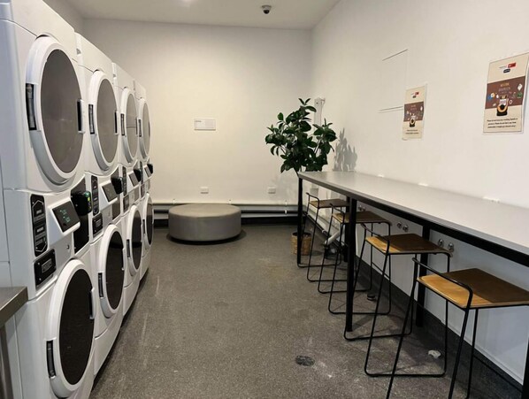 A laundry room with multiple washing machines, a seating area with stools, and a potted plant. - Essence Hotel Carlton (Carlton)