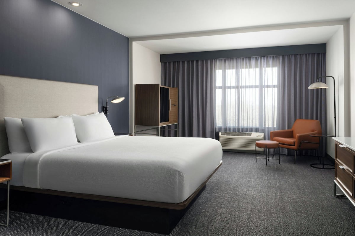 A modern hotel room with a large bed, a desk, an armchair, and a window with curtains.