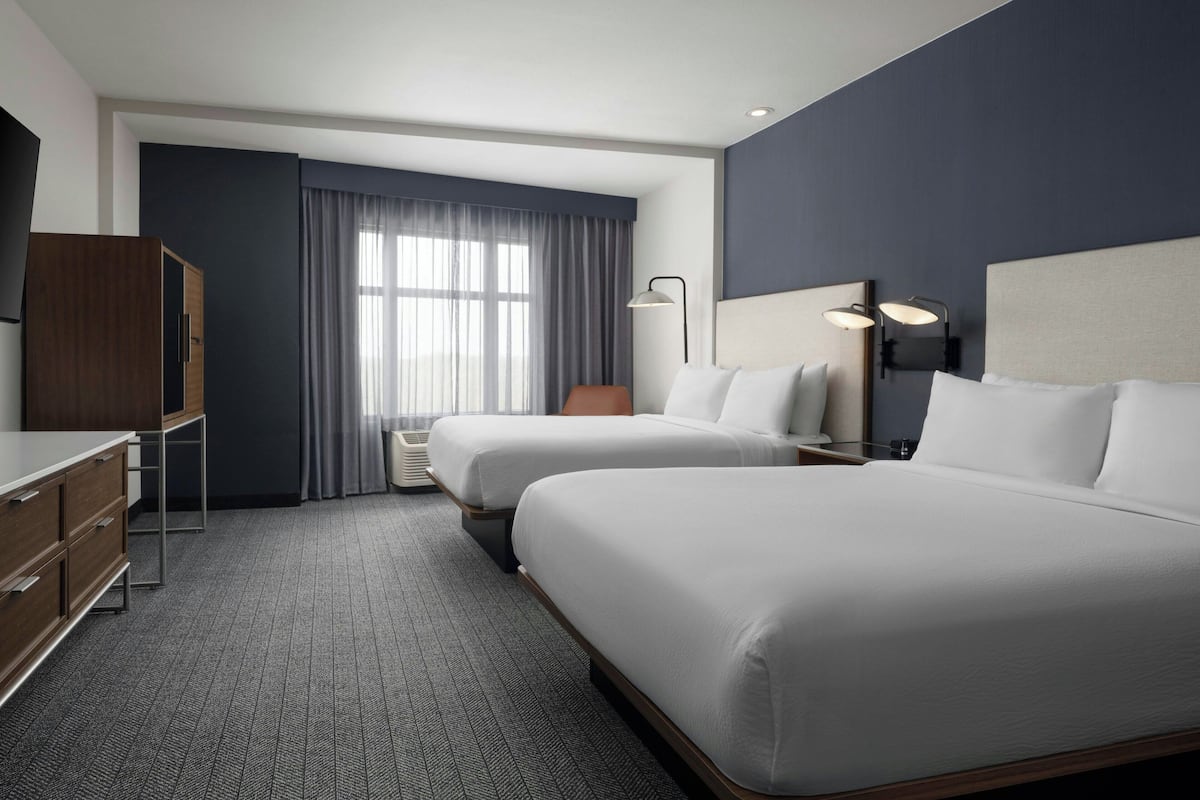 A hotel room with two beds, a desk, a wardrobe, and a large window with curtains.