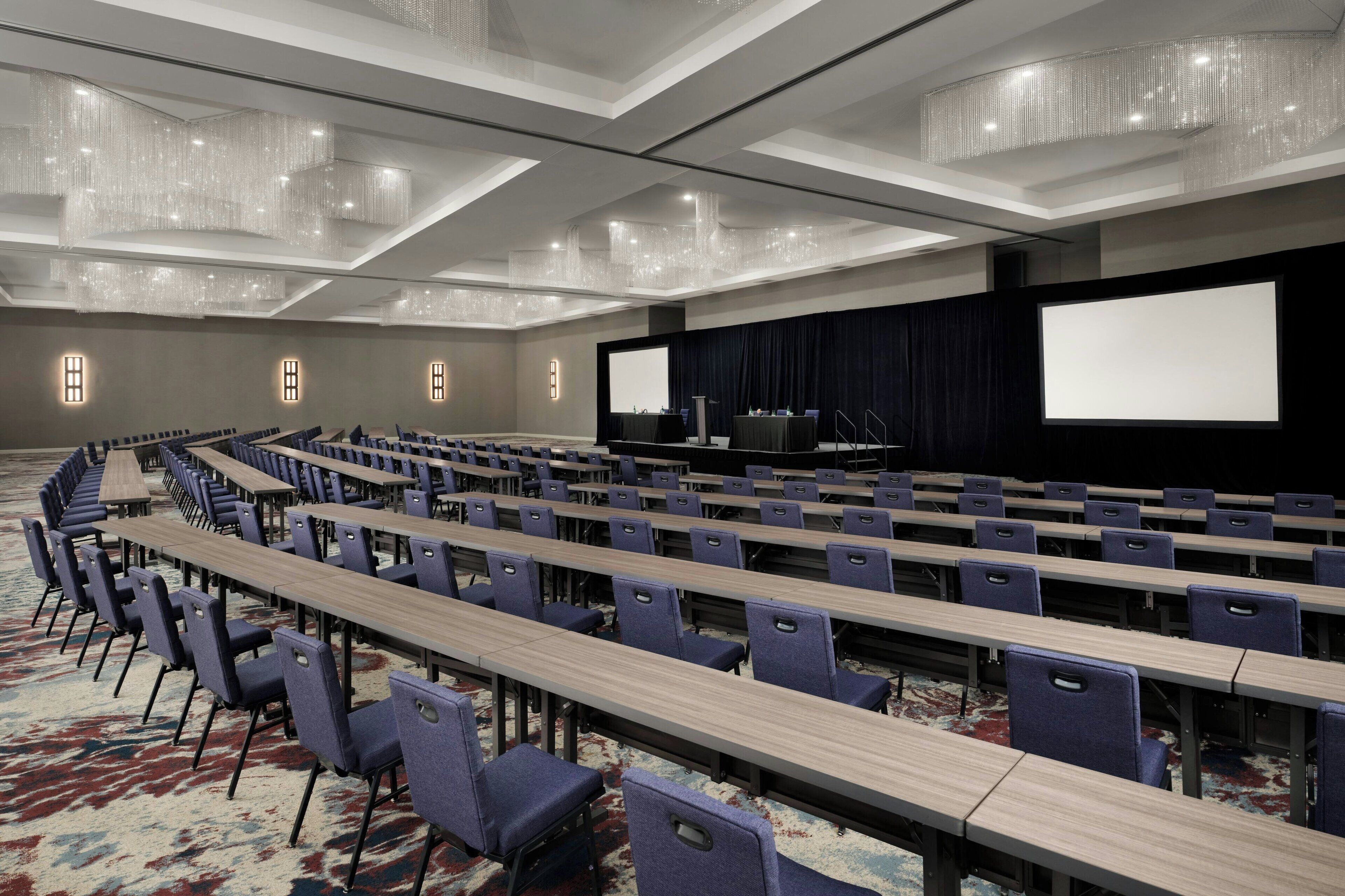 A conference room with rows of tables and chairs, a large screen, and a stage.