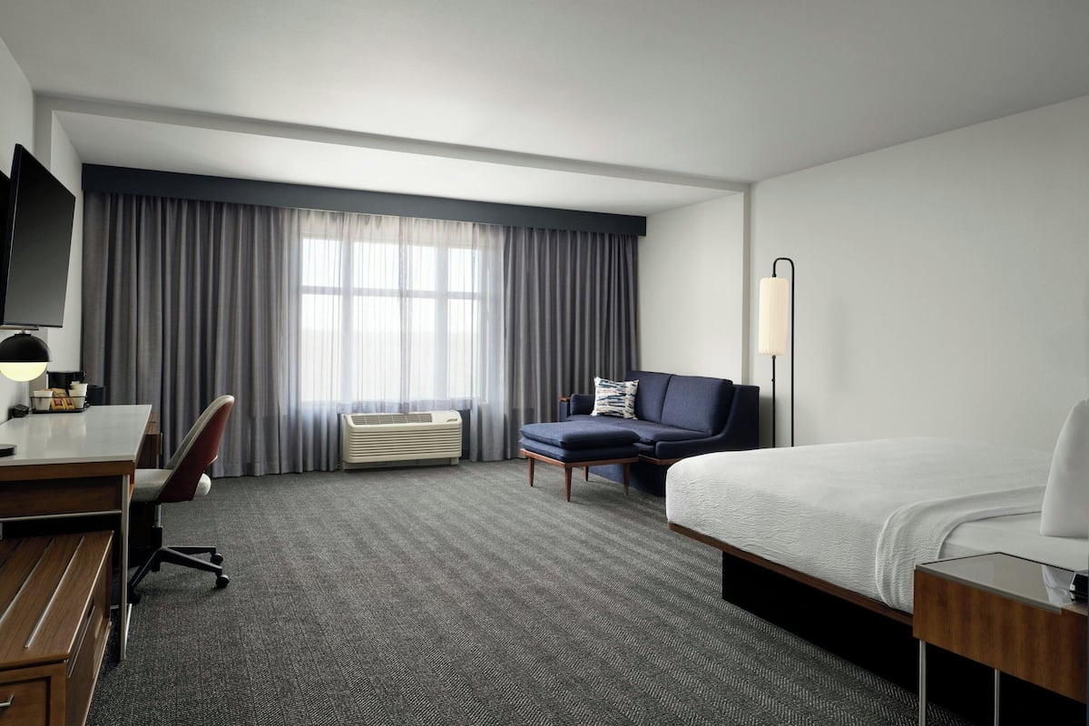 A hotel room with a bed, a desk with a chair, a sofa, a lamp, and a large window with curtains.