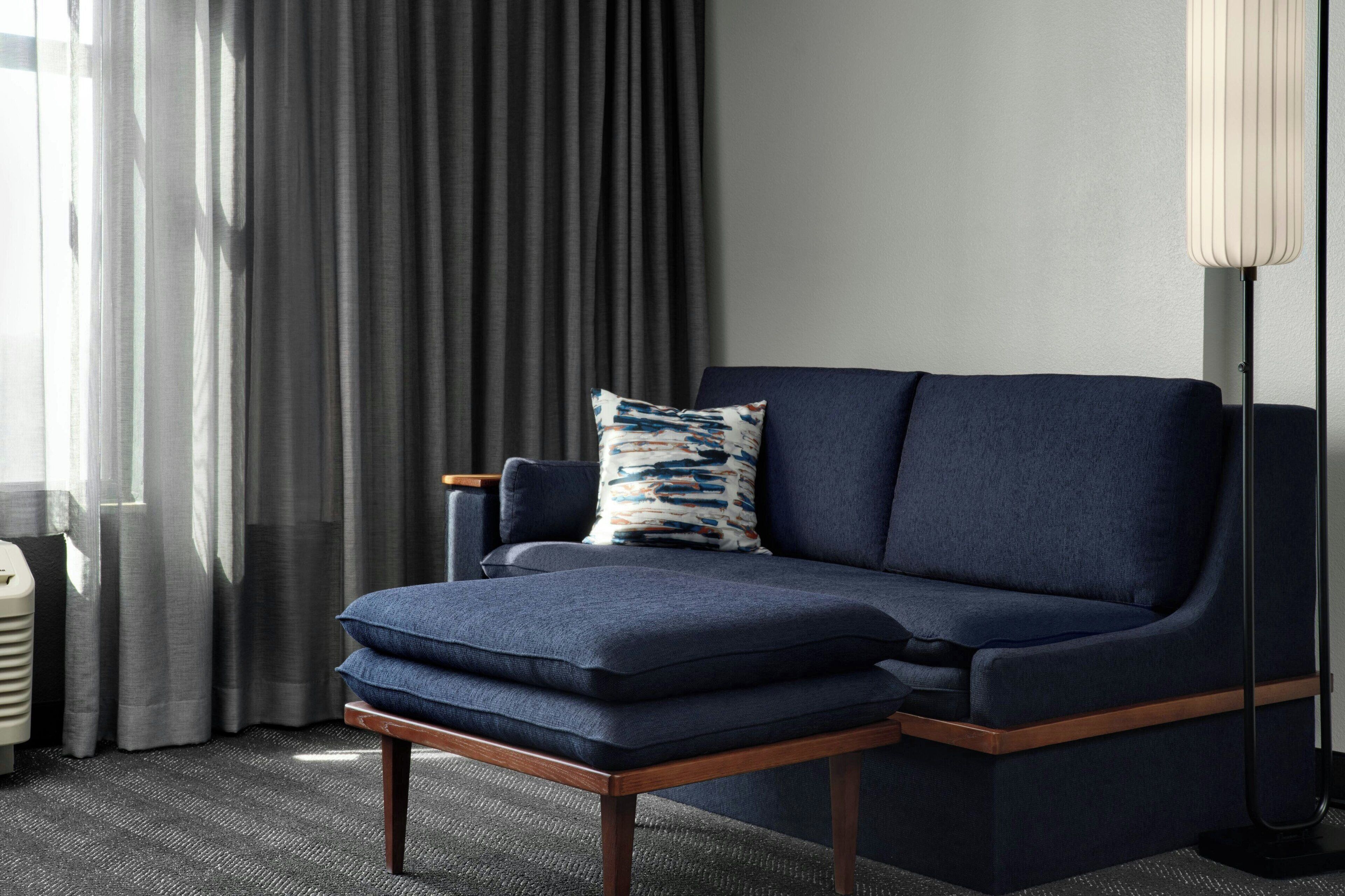 A modern living room with a dark blue sofa, a matching ottoman, and a patterned throw pillow. A floor lamp is positioned to the right, and a radiator is visible on the left.