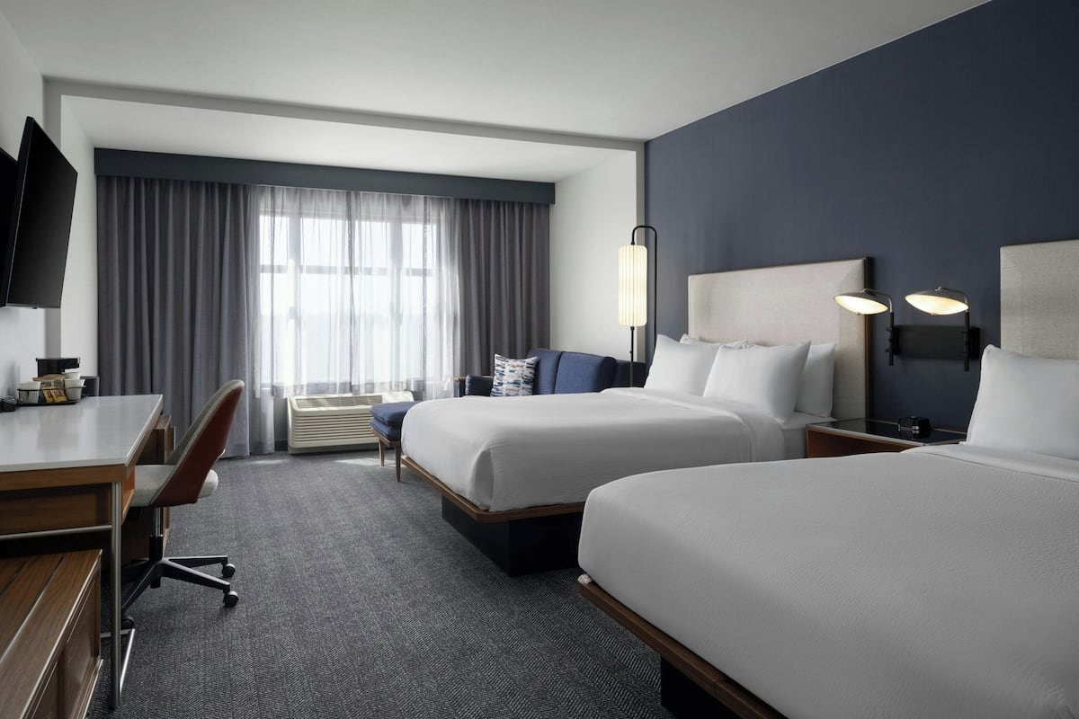 A hotel room with two beds, a desk, a chair, a TV, and a large window with curtains.