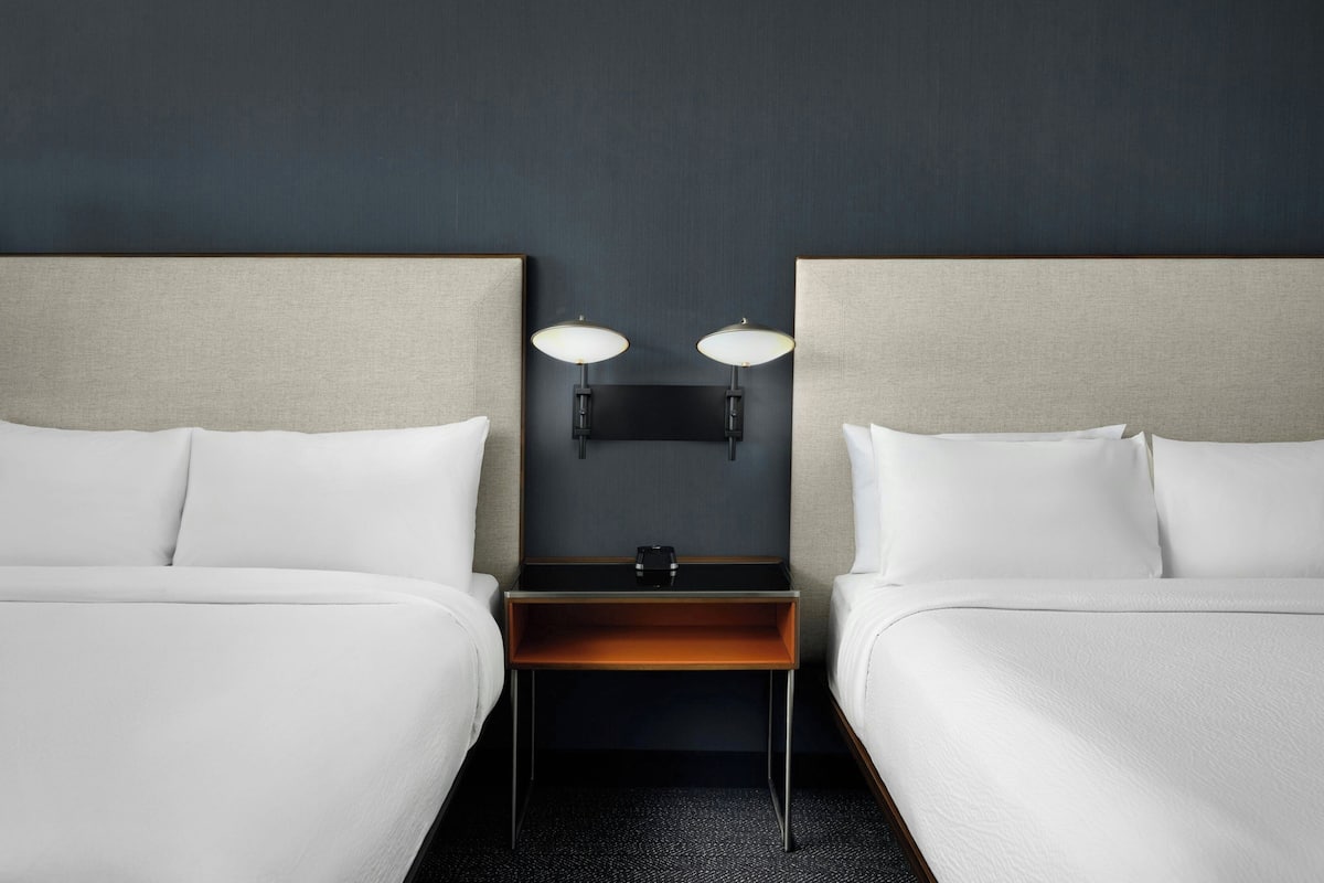 A hotel room with two beds, a nightstand with a lamp, and a flat-screen TV mounted on the wall.