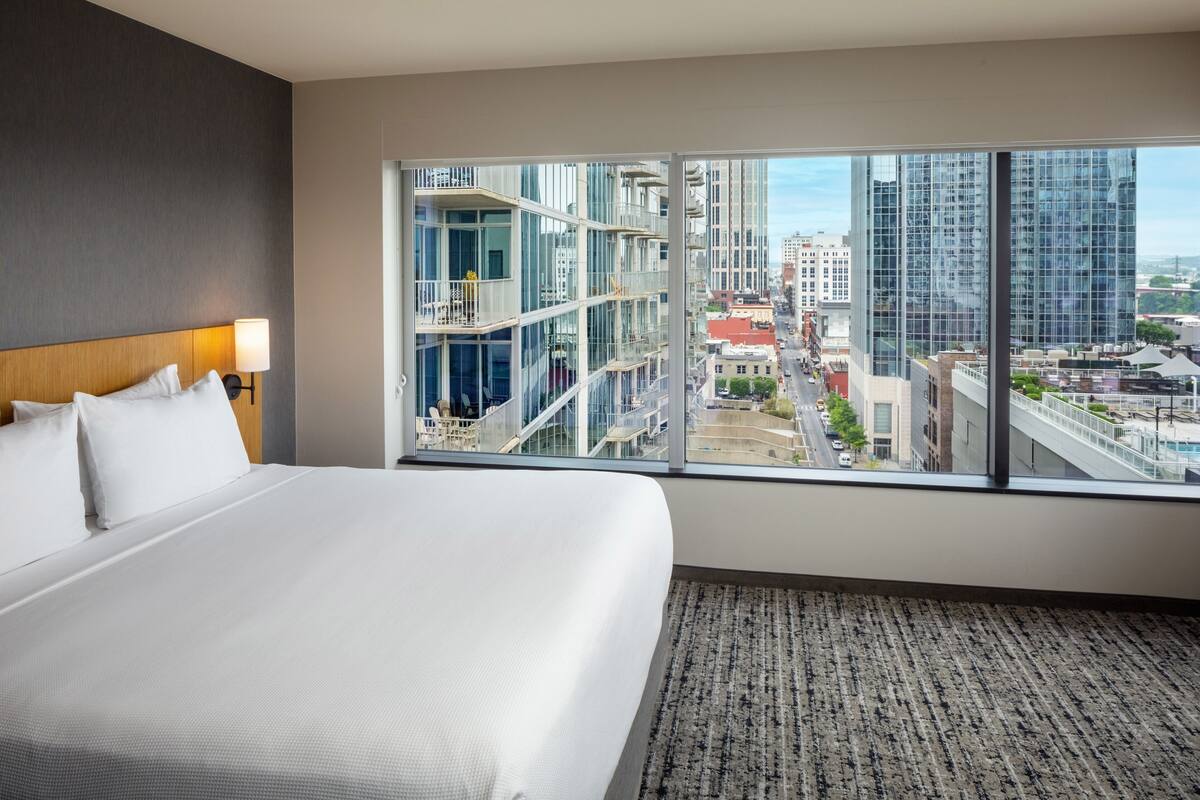 Habitación de hotel con una cama grande, vistas de la ciudad a través de ventanales del suelo al techo y un diseño moderno.
