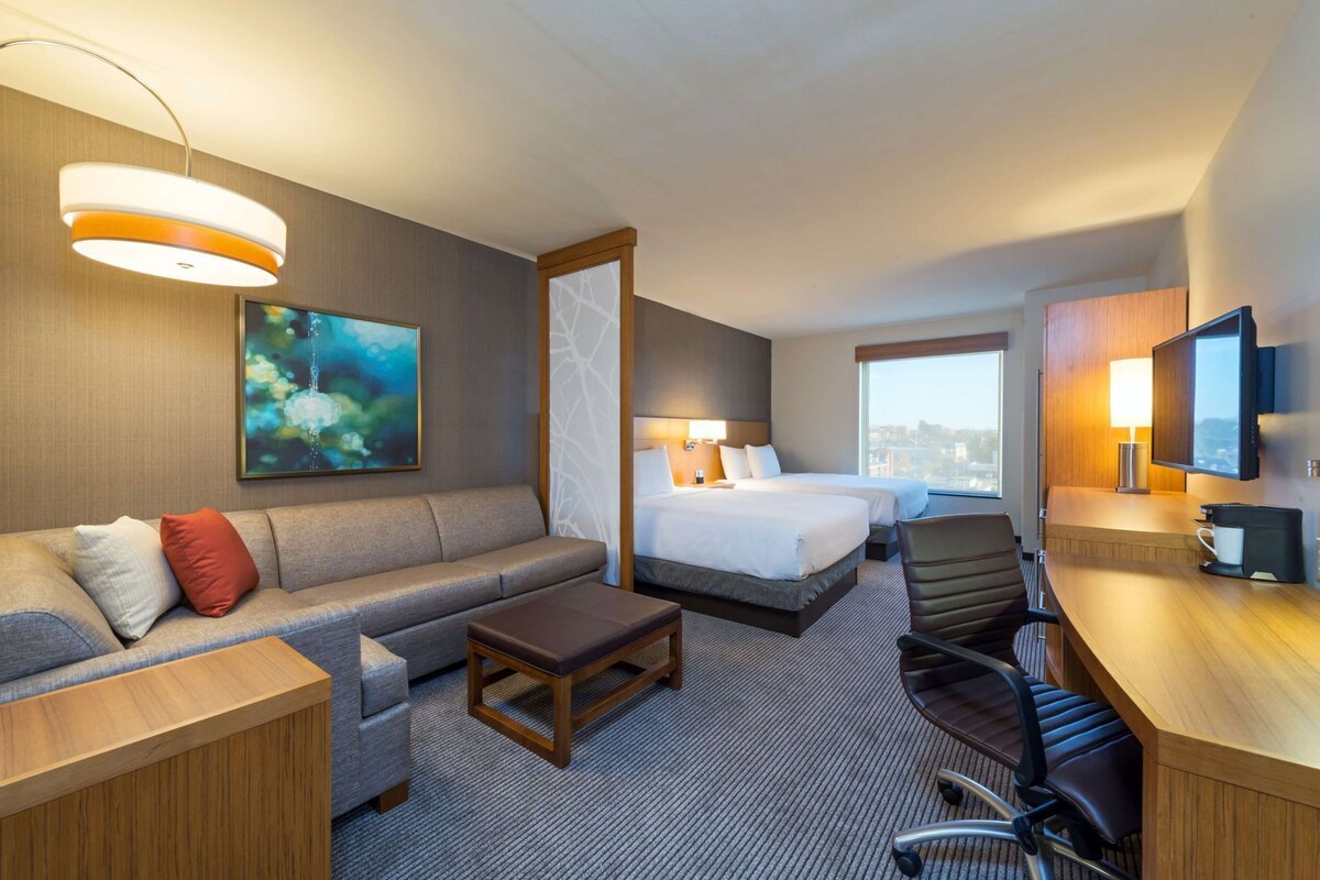 Ein Hotelzimmer mit einer Couch, zwei Betten, einem Schreibtisch, einem Sessel, einem Fernseher und einem großen Fenster.