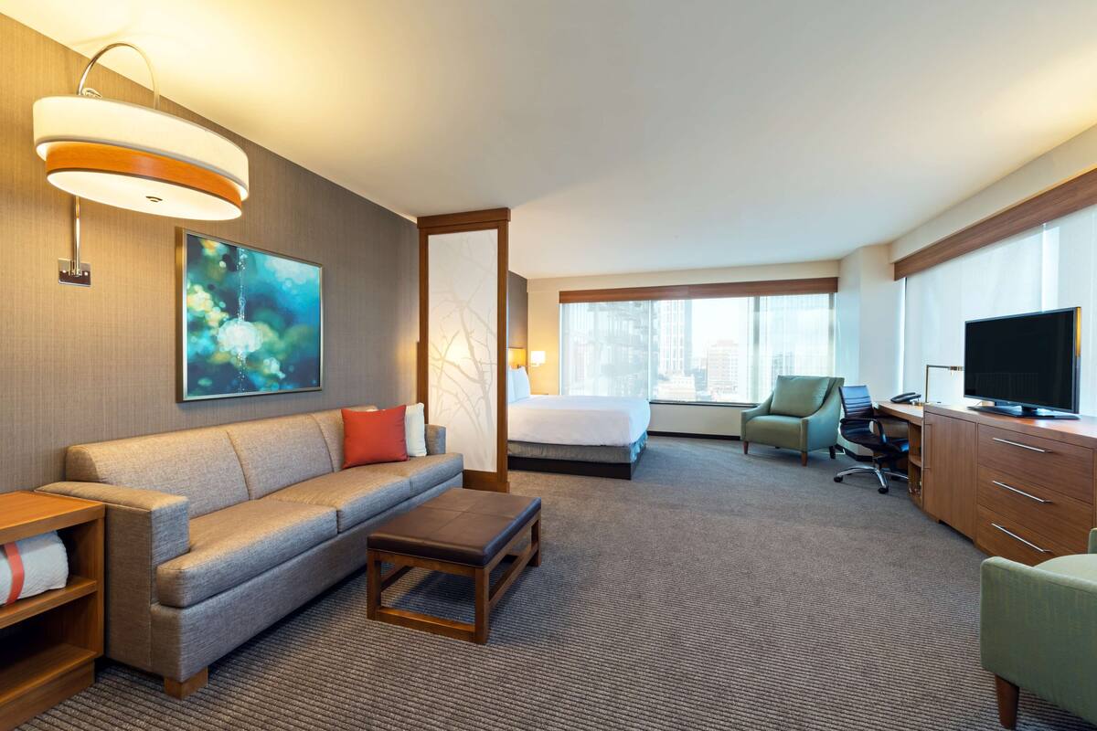 Habitación de hotel con sofá, cama, escritorio con televisor y ventana con vista a la ciudad.