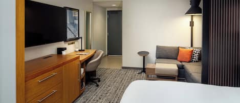 Una habitación de hotel con una cama, un escritorio con silla, televisor montado en la pared, una lámpara y un sofá con almohadas.