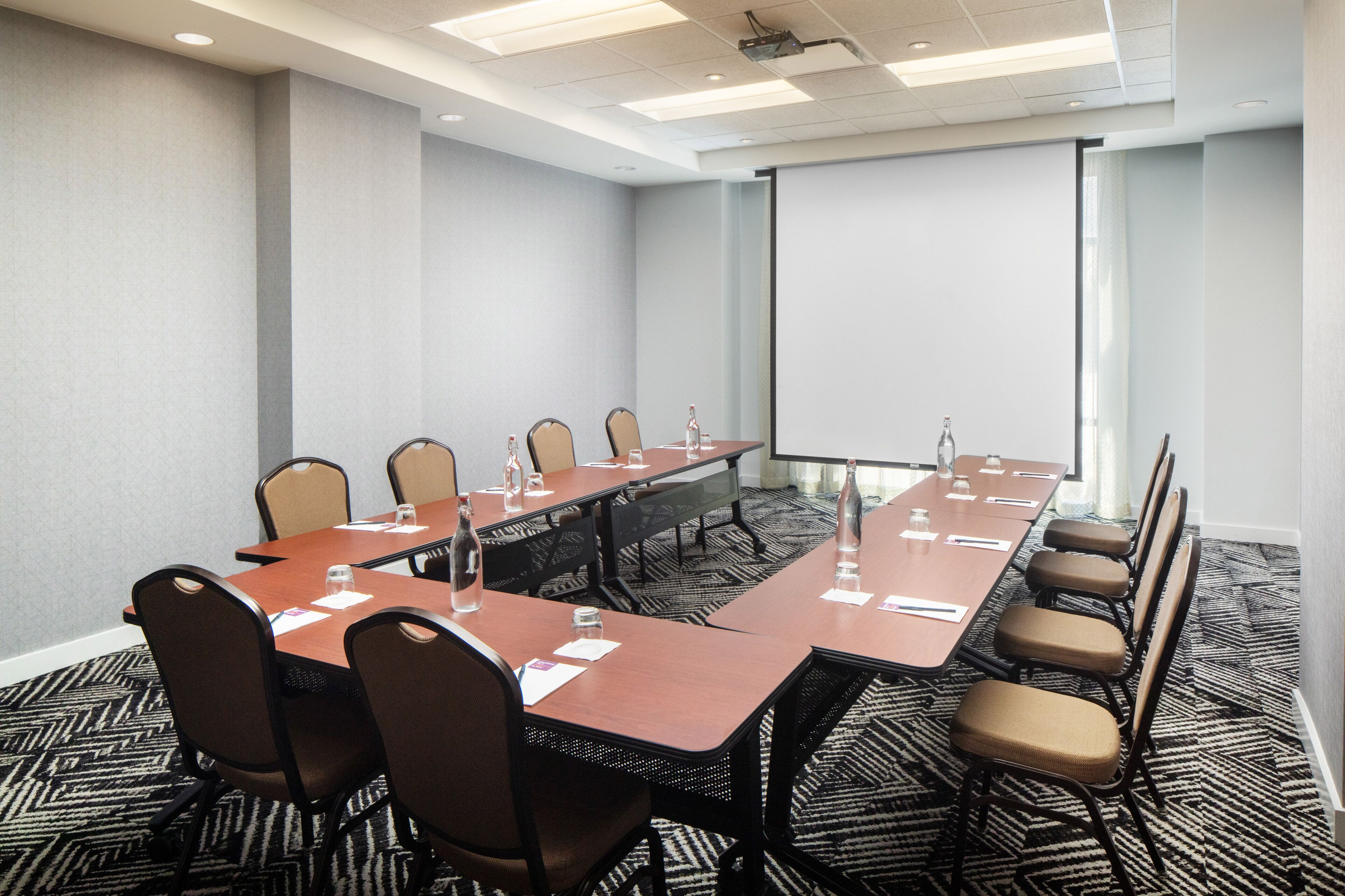 Un salón de conferencias con una larga mesa, sillas, una pantalla de proyector y piso alfombrado.