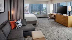 Een hotelkamer met een bed, bank, bureau, televisie en een groot raam met uitzicht op de stad.