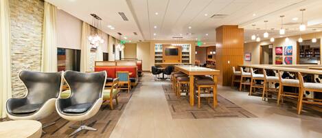 A modern hotel lobby with a central seating area, a bar, and a TV screen.