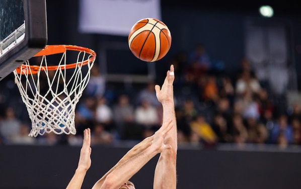 Una canasta de baloncesto con red, un balón en suspensión y un público desenfocado al fondo.